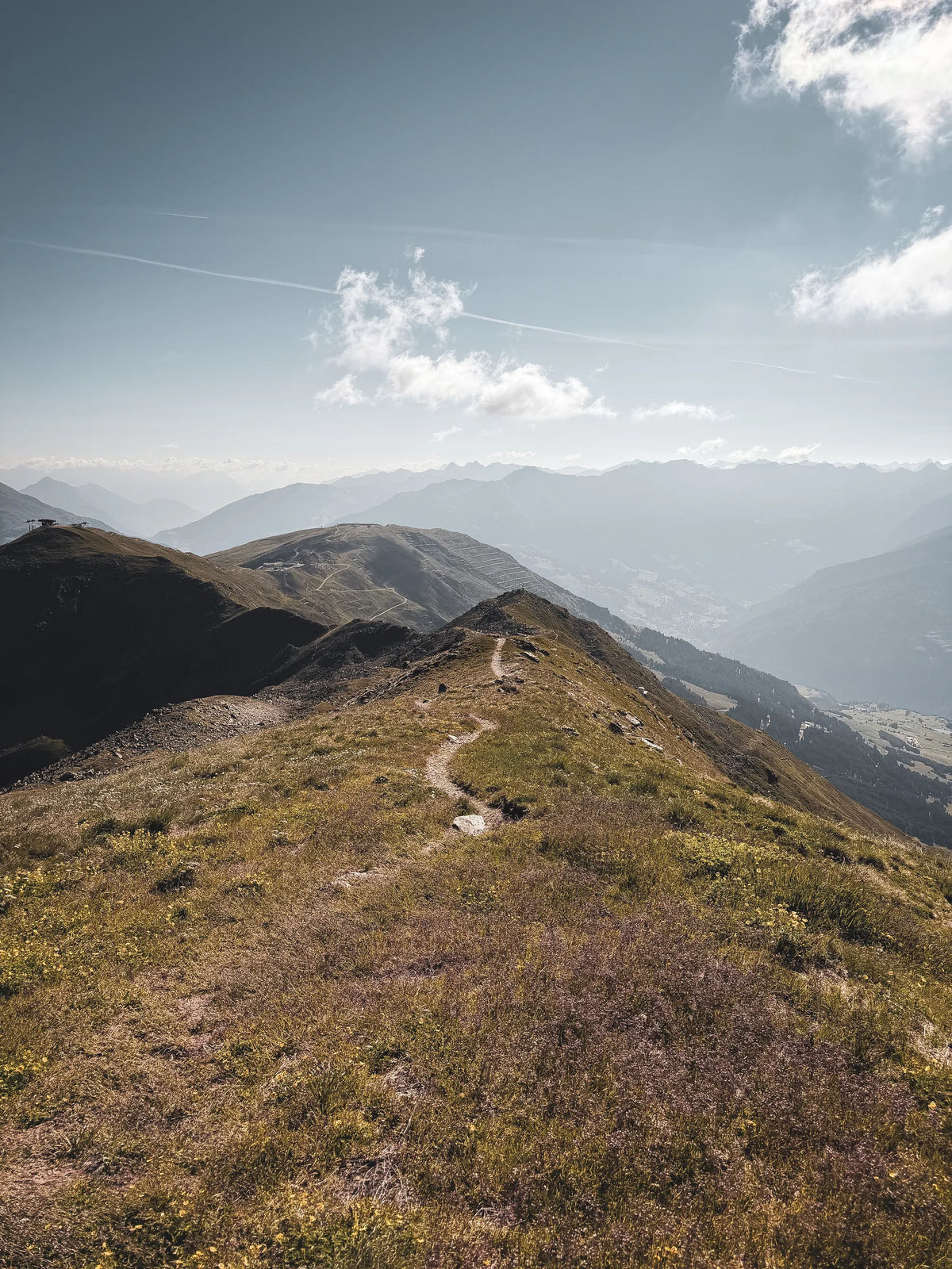 Schmaler Pfad über einen grasbewachsenen Grat, dahinter Bergketten im Dunst